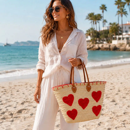 Panier en raphia beige naturel à cœurs rouges et longues anses marron, porté à la main sur la plage.
