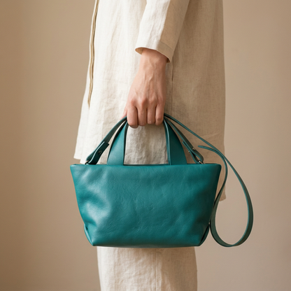 Sac Juanita en cuir turquoise, tenu à la main par ses poignées sur fond beige, Bmassy.