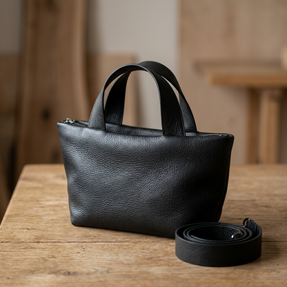 Sac Juanita en cuir noir, posé sur une table en bois avec sa bandoulière enroulée à côté, Bmassy.