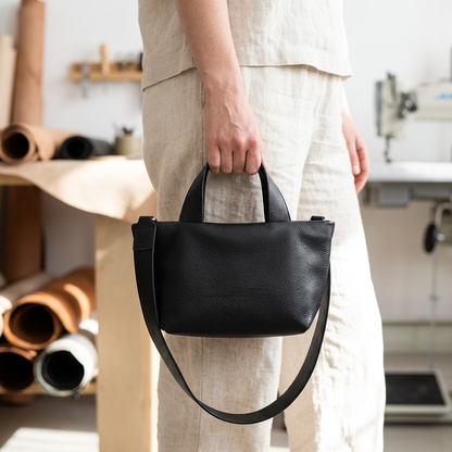Sac Juanita en cuir noir, tenu à la main par ses poignées dans un atelier, Bmassy.