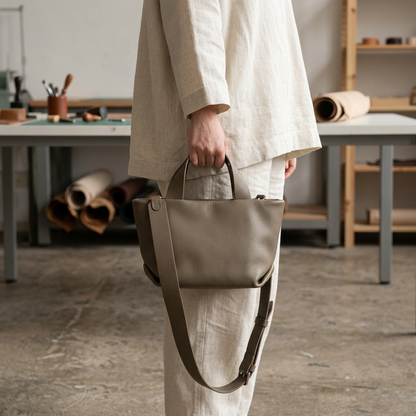 Sac Juanita en cuir gris, tenu à la main par ses poignées devant une tenue écrue, Bmassy.