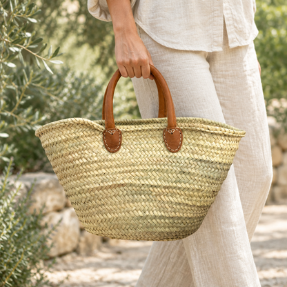 Cabas en raphia beige naturel à anses en cuir camel, porté à la main dans un jardin