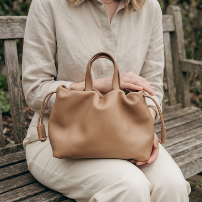 Petit sac à main Angelines en cuir beige sable posé sur les genoux d’une femme assise sur un banc, Bmassy.