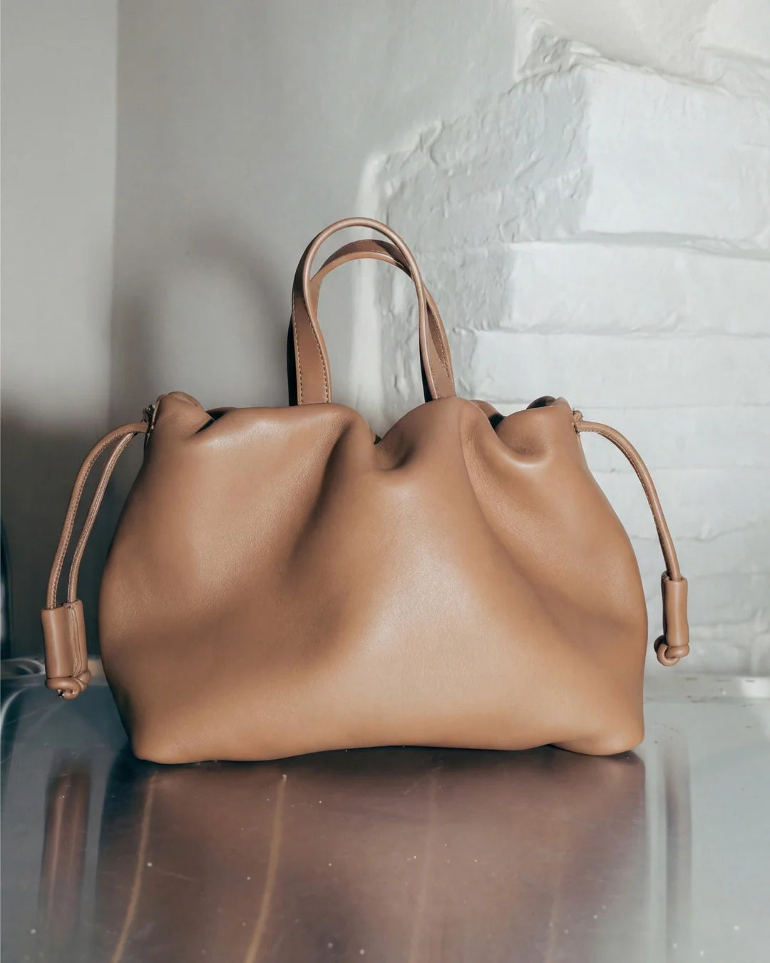 Petit sac à main Angelines en cuir beige sable vu de face avec ses poignées et ses cordons de serrage, posé sur une surface réfléchissante devant un mur blanc, Bmassy.