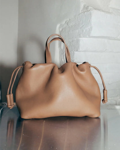 Petit sac à main Angelines en cuir beige sable vu de face avec ses poignées et ses cordons de serrage, posé sur une surface réfléchissante devant un mur blanc, Bmassy.