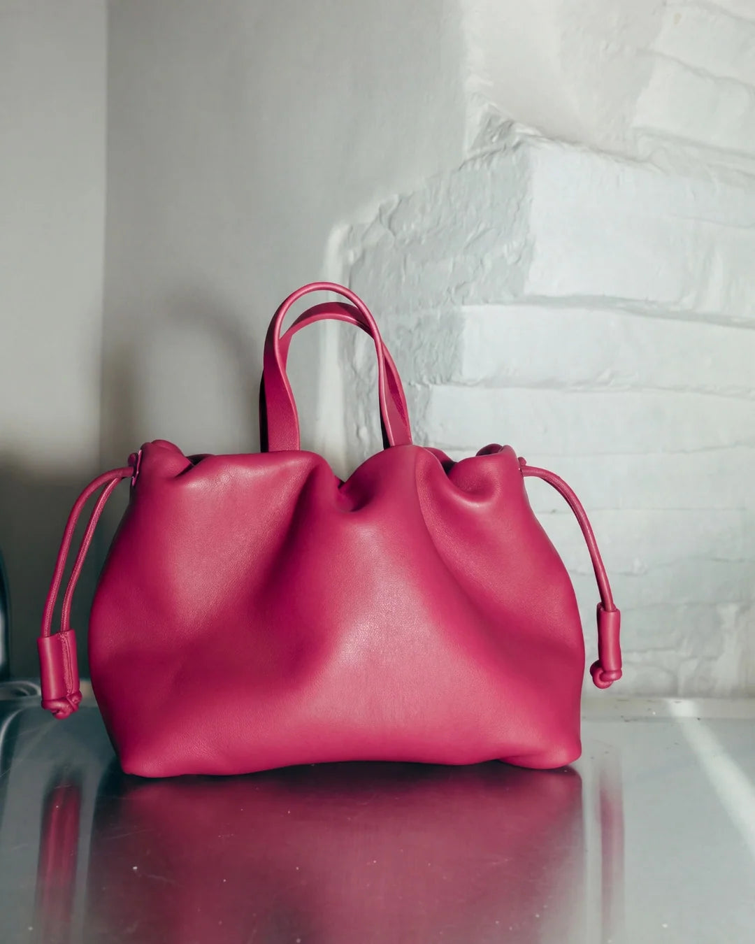 Petit sac à main Angelines en cuir rose framboise vu de face avec ses poignées et ses cordons de serrage devant un mur blanc, Bmassy.