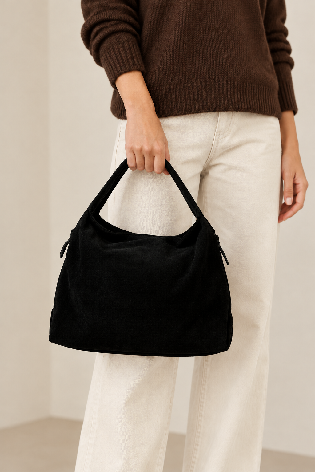 Sac Capucine en cuir velours noir porté à la main, avec un pull brun et un pantalon écru