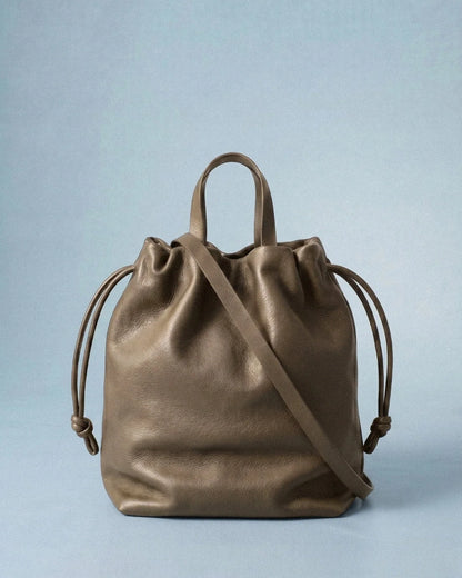 Sac à main Angela Medium en cuir gris taupe vu de face avec sa bandoulière et ses cordons de serrage sur fond bleu clair, Bmassy.
