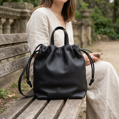 Sac à main Angela Medium en cuir noir posé sur les genoux d’une femme assise sur un banc, Bmassy.