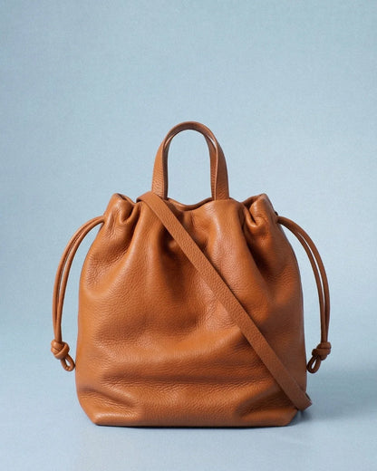 Sac à main Angela Medium en cuir orange cuivré vu de face avec sa bandoulière et ses cordons de serrage sur fond bleu clair, Bmassy.
