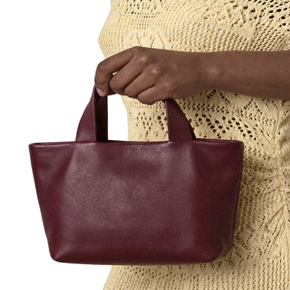 Sac Juanita en cuir bordeaux, tenu à la main par ses poignées devant une tenue écrue, Bmassy.
