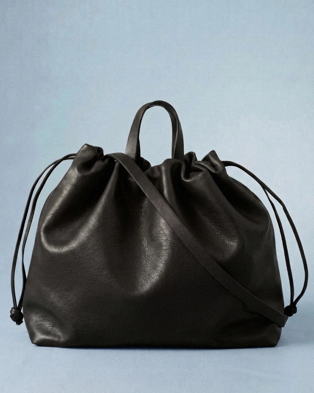 Sac à main maxi Dona Angela en cuir noir vu de face, avec ses poignées, sa bandoulière et ses cordons de serrage sur fond bleu clair, Bmassy.