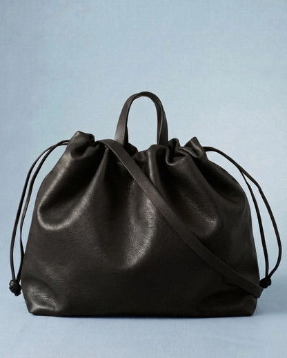 Sac à main maxi Dona Angela en cuir noir vu de face, avec ses poignées, sa bandoulière et ses cordons de serrage sur fond bleu clair, Bmassy.