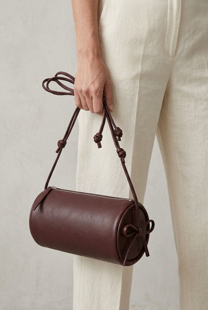 Sac Alma bordeaux porté à la main par ses longs cordons noués, sur un pantalon écru
