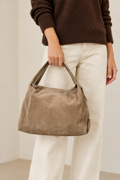 Sac Capucine en cuir velours taupe noisette porté à la main, avec un pull brun et un pantalon écru
