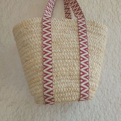 Cabas en raphia beige naturel, avec longues anses à motifs géométriques bordeaux et écrus, suspendu contre un mur.